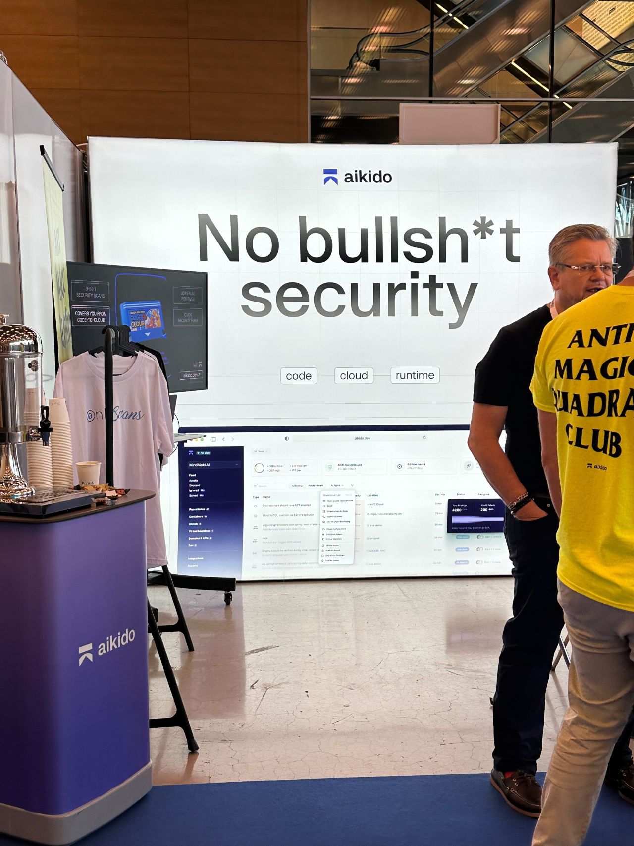 Exhibition booth with a large sign that reads 'No bullsh*t security' and two men talking, one wearing a yellow shirt with 'ANTI MAGIC QUADRA CLUB' text.