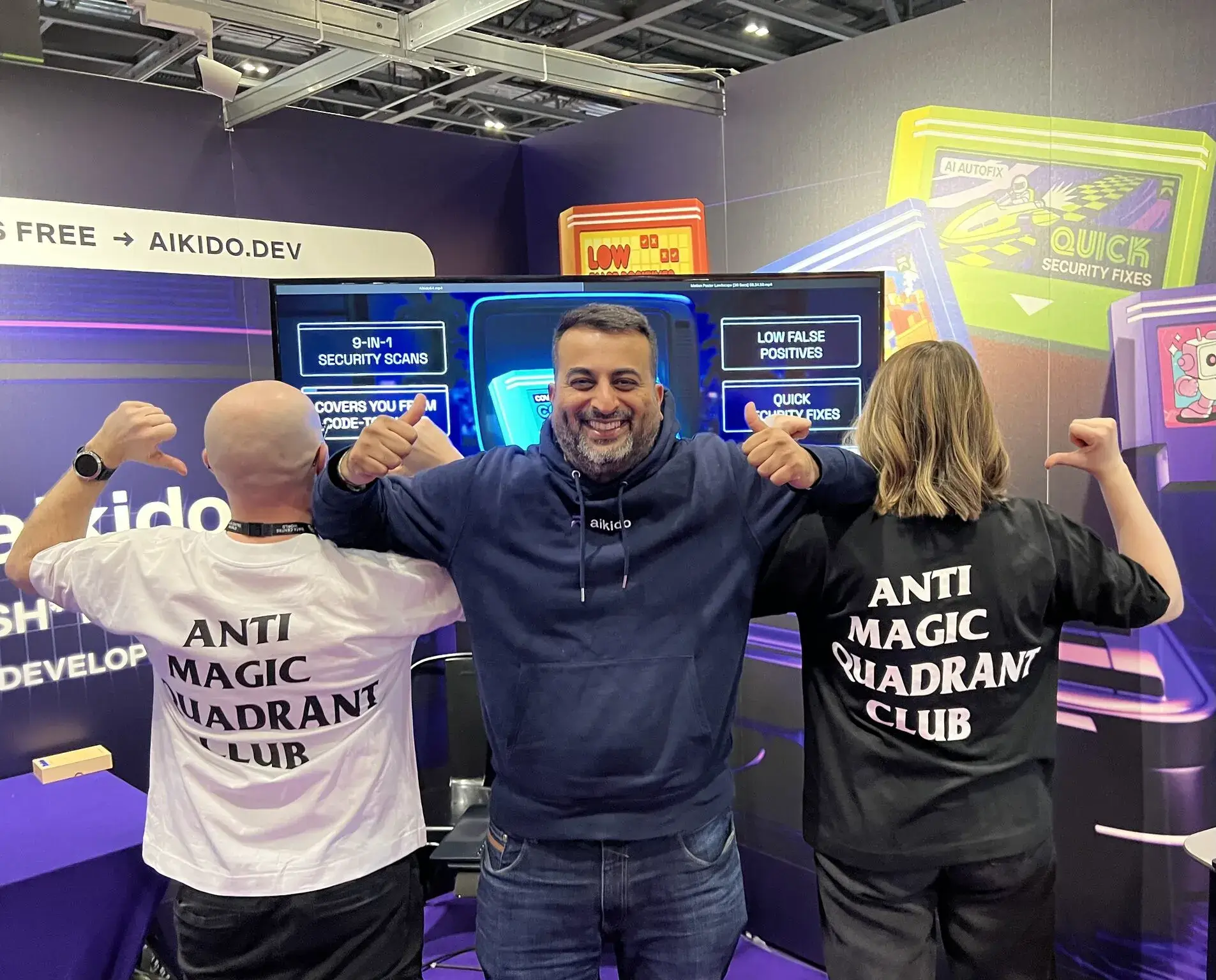 A man in a navy hoodie stands smiling between a bald man and a woman, both pointing thumbs at the text 'ANTI MAGIC QUADRANT CLUB' on their white and black t-shirts.