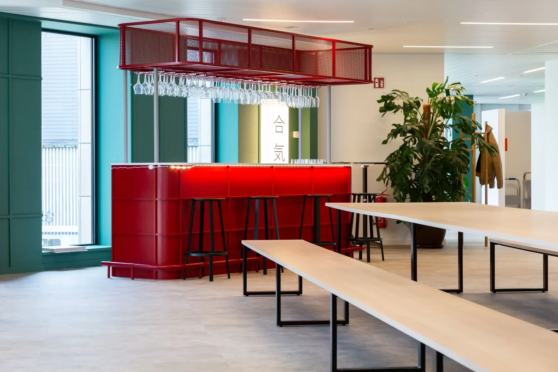Modern indoor space with a red bar counter, black stools, hanging wine glasses, large windows, light wooden tables, and green plants.