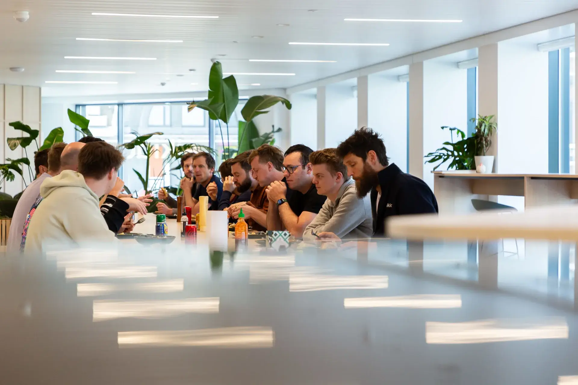Group of men sitting in a modern office dining area, eating and talking around a long table with plants and condiments.