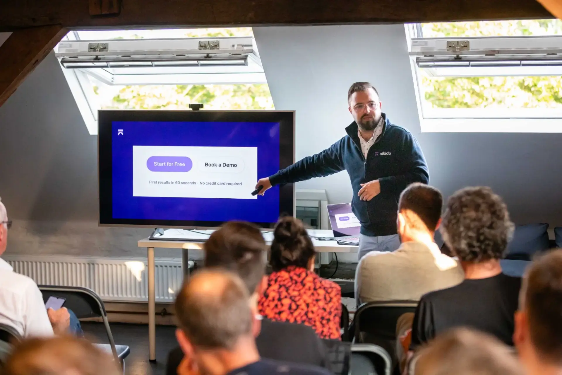 Man in glasses and dark jacket giving a presentation to a seated audience, pointing at a screen displaying 'Start for Free' and 'Book a Demo' buttons.