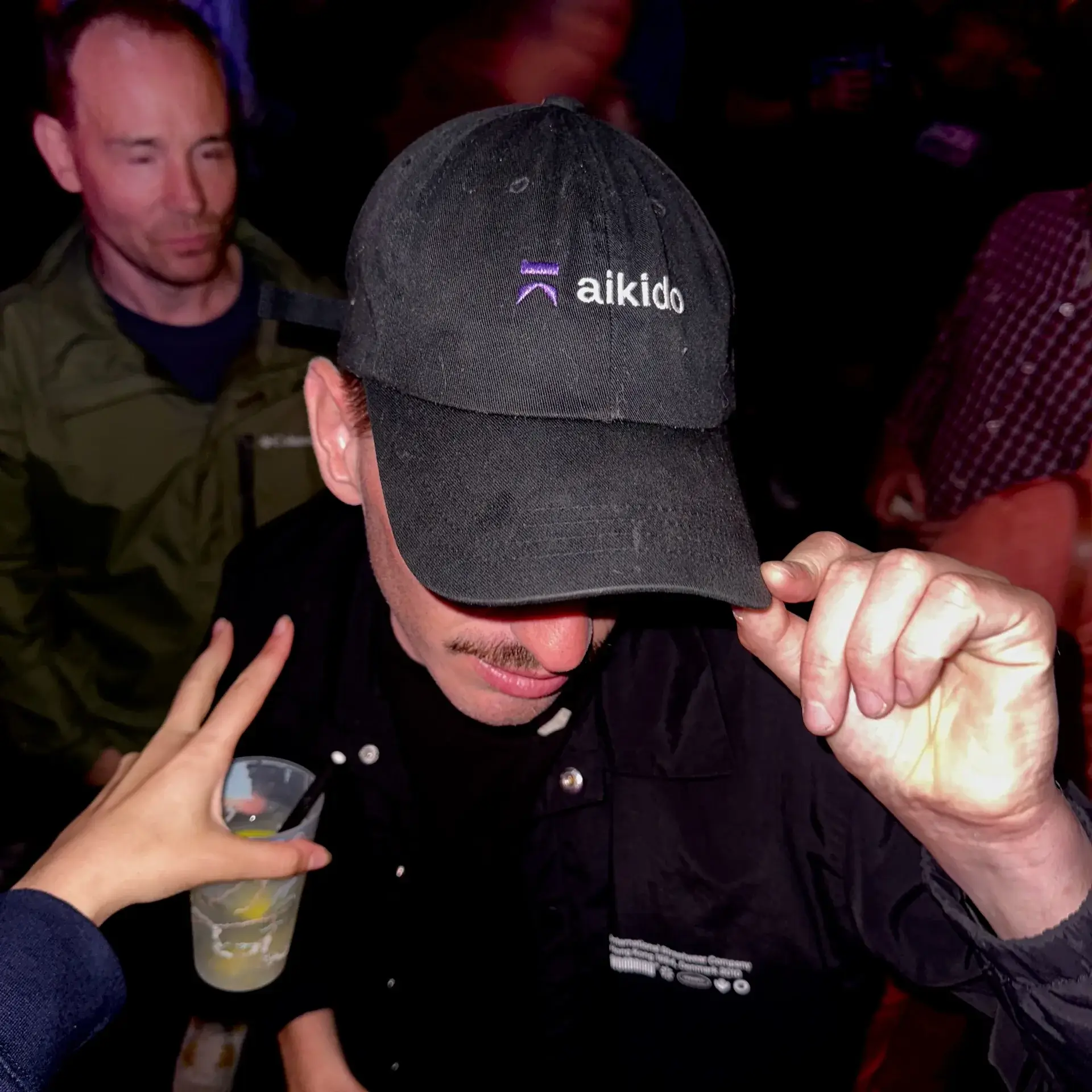 Person wearing a black cap with 'aikido' logo, tilting the cap down while another hand holds a cup with a straw nearby.