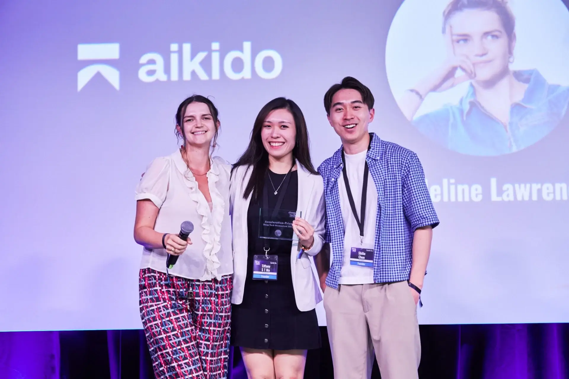 Three smiling people standing on stage in front of a screen with the aikido logo, one holding a microphone and another holding an award plaque.