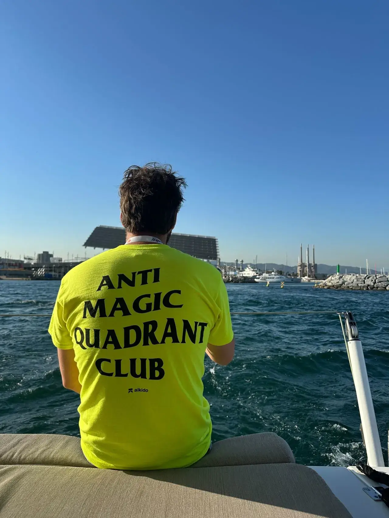 Man wearing a bright yellow shirt with text 'ANTI MAGIC QUADRANT CLUB' sitting on the edge of a boat facing the water and marina.