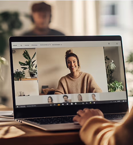 Laptop screen displaying a video call with a smiling woman in a beige sweater, blurred person's hand in foreground.