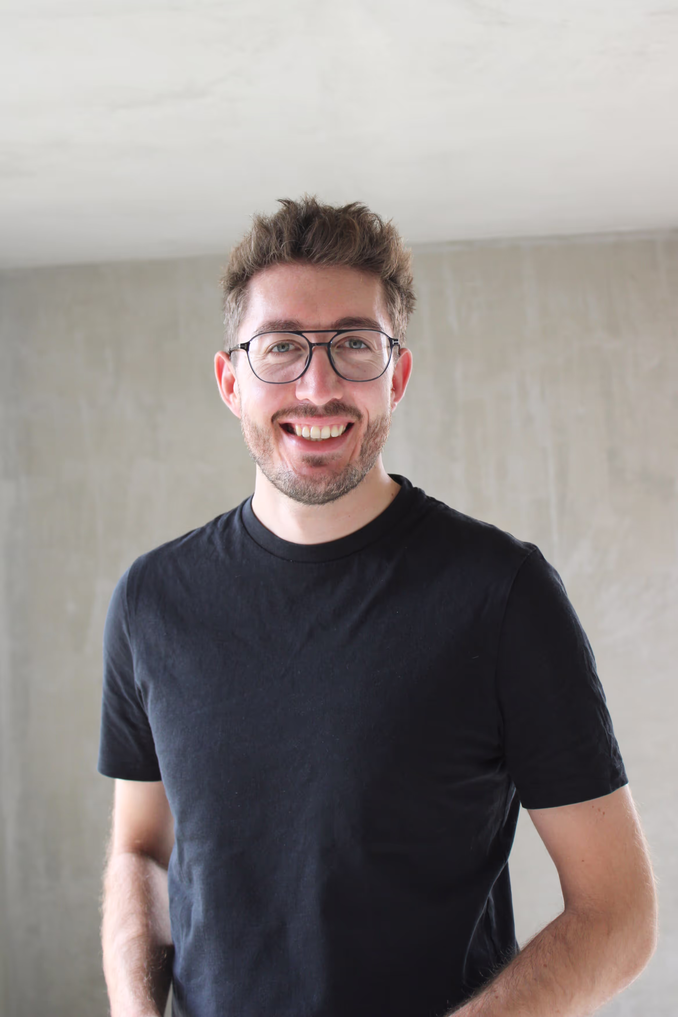 Lächelnder Mann mit Brille und schwarzem T-Shirt vor grauer Wand