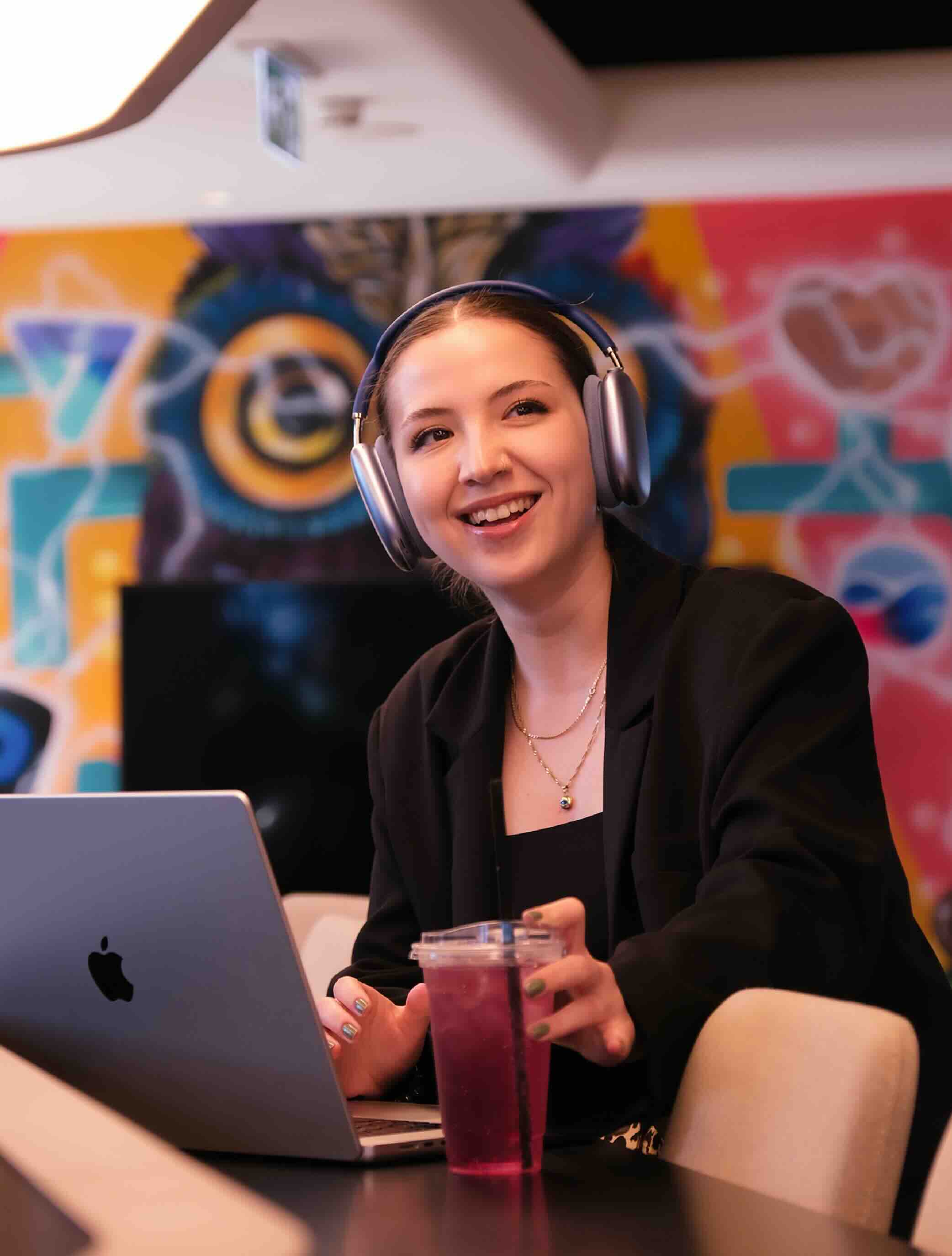 Girl in front of her laptop. She has headphones on her head and a drink in her left hand. She's smiling and looking slightly behind the camera.