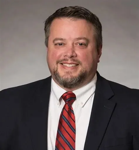 Smiling man with short dark hair and beard wearing a dark suit jacket, white shirt, and red striped tie.