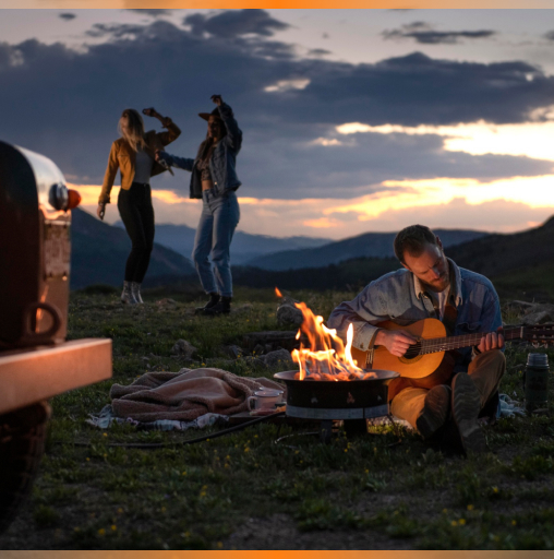 Man playing guitar by a campfire while two women dance in the background during sunset in a mountainous area.