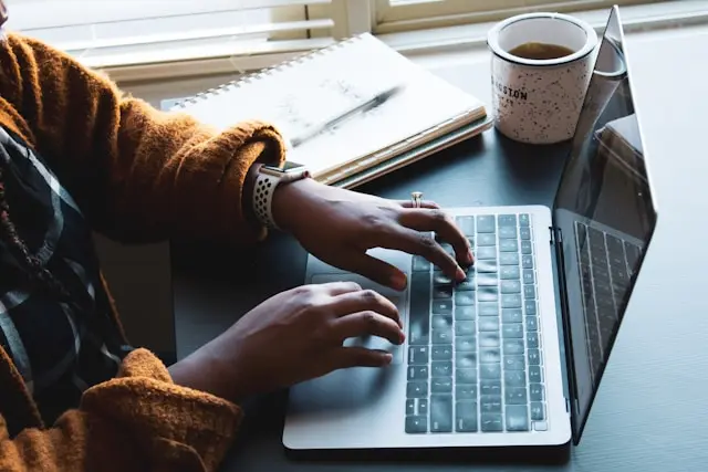 Person typing on a laptop at a desk with a notebook and coffee nearby.