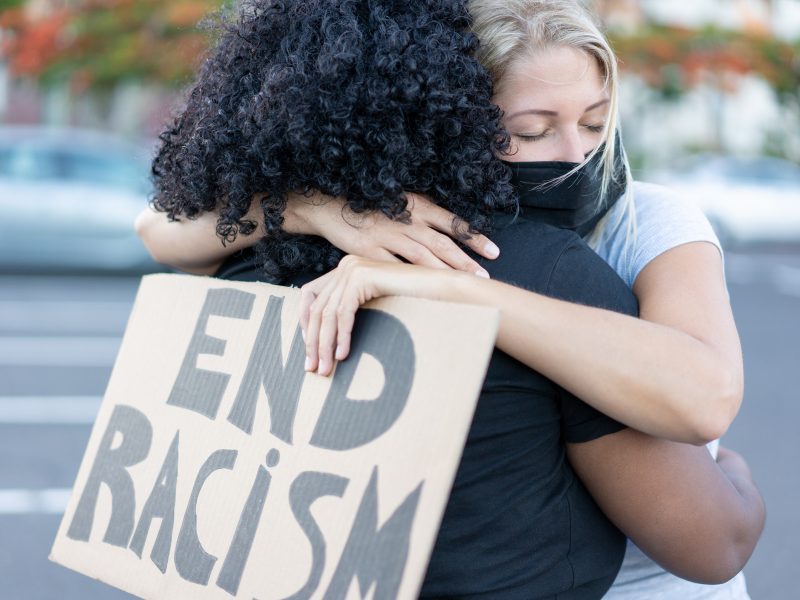 Two individuals embrace while holding a sign that reads "END RACISM," symbolizing solidarity in anti-racism efforts.