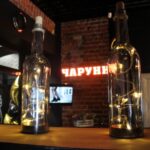 На полке выставлены три стеклянные бутылки с пробками, каждая из которых украшена гирляндами. На заднем плане - кирпичная стена с неоновой вывеской и телевизионным экраном. Судя по всему, обстановка в помещении, возможно, в баре или кафе. На полке выставлены три стеклянные бутылки с пробками, каждая из которых украшена гирляндами. На заднем плане - кирпичная стена с неоновой вывеской и телевизионным экраном. Судя по всему, обстановка в помещении, возможно, в баре или кафе.