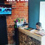 Женщина сидит за деревянной стойкой ресепшн, сложенной из дров, и разговаривает по телефону. Телевизор на кирпичной стене показывает танцоров, а над ним светится неоновая вывеска с кириллическими буквами. На столе лежат цветы и книги. Женщина сидит за деревянной стойкой ресепшн, сложенной из дров, и разговаривает по телефону. Телевизор на кирпичной стене показывает танцоров, а над ним светится неоновая вывеска с кириллическими буквами. На столе лежат цветы и книги.