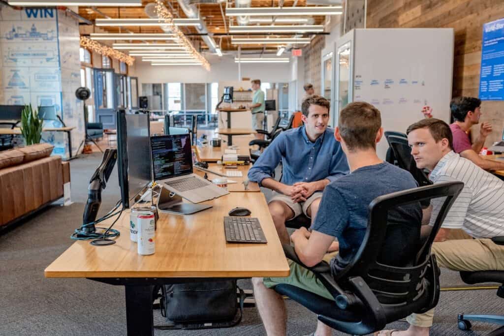 Three men seated in a room having a conversation at a desk with a computer and drinks on it salary after a coding bootcamp