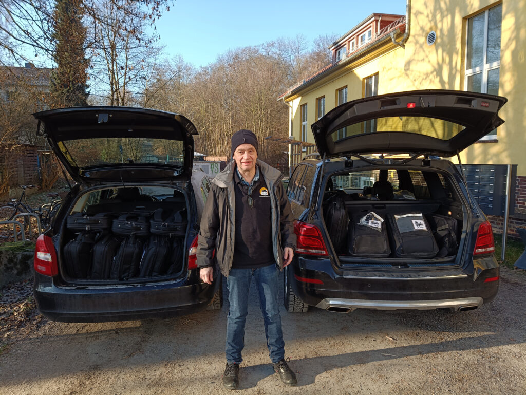 Andreas steht im Freien zwischen zwei Autos, bei denen die Kofferraumdeckel geöffnet sind. Im Inneren beider Fahrzeuge befinden sich jede Menge Laptoprucksäcke.