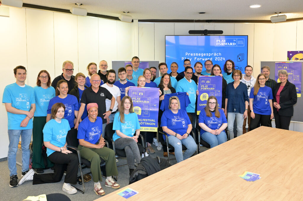 Das Foto zeigt ein Gruppenbild mit ca. drei Dutzend Personen in einem Konferenzraum. Viele tragen ein blaues T-Shirt mit dem aktuellen „Play Forward“-Logo. Zwei Personen halten das Veranstaltungsplakat in die Kamera. Auf einem großen Bildschirm im Hintergrund wird der Text „Pressegespräch Play Forward 2024“ angezeigt.