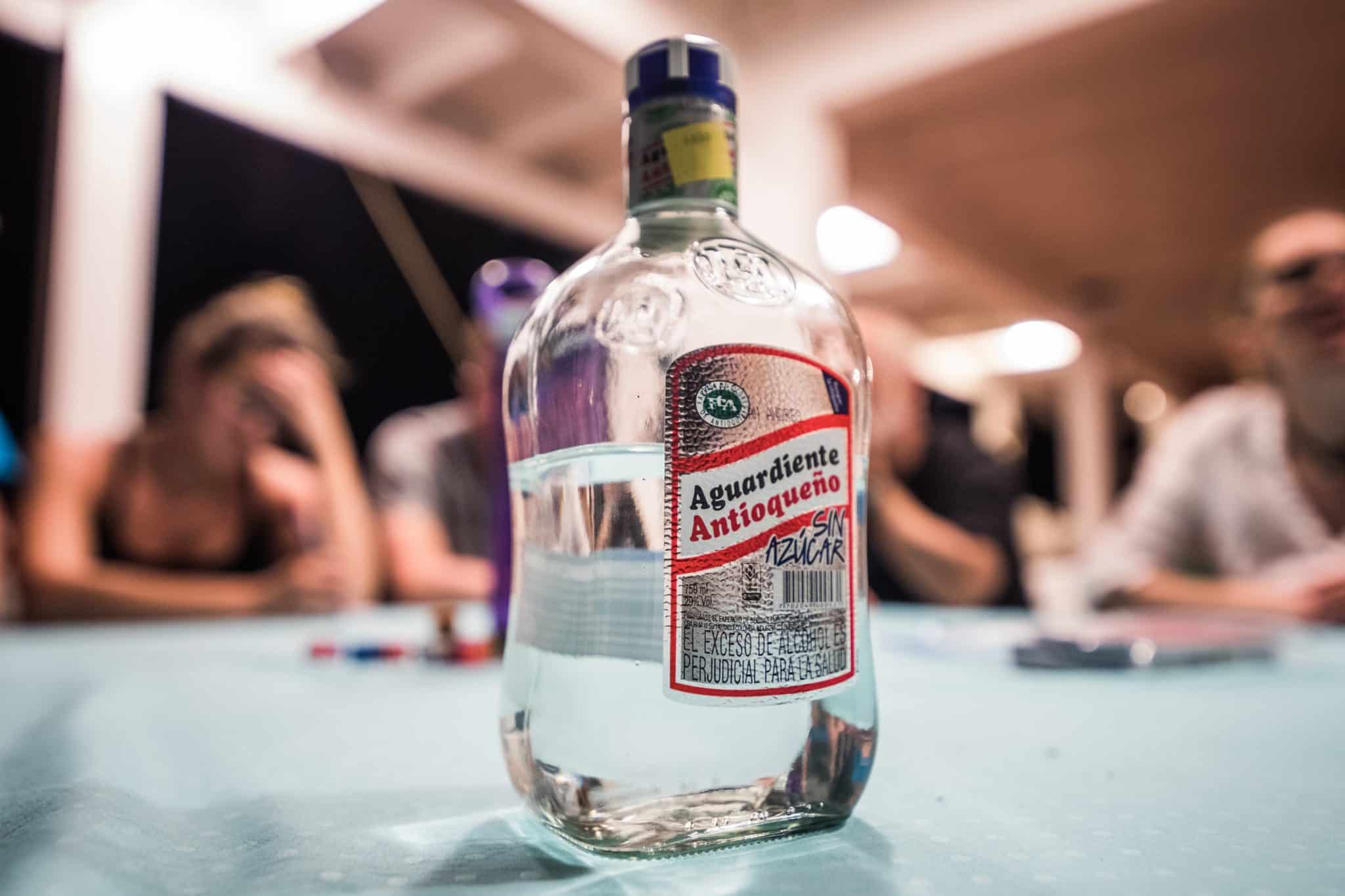 A clear bottle of Aguardiente Antioqueño on a table, with blurry people in the background, suggesting a social setting.