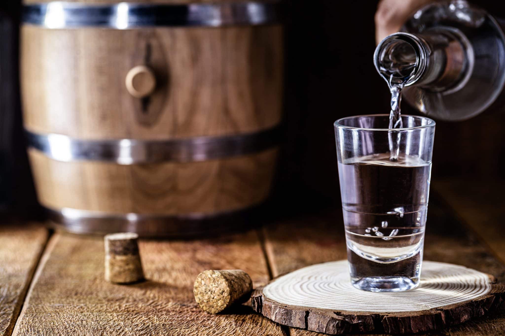 Liquid is being poured into a clear glass from a bottle, with a wooden barrel in the background, all set on a wooden surface with a rustic feel.