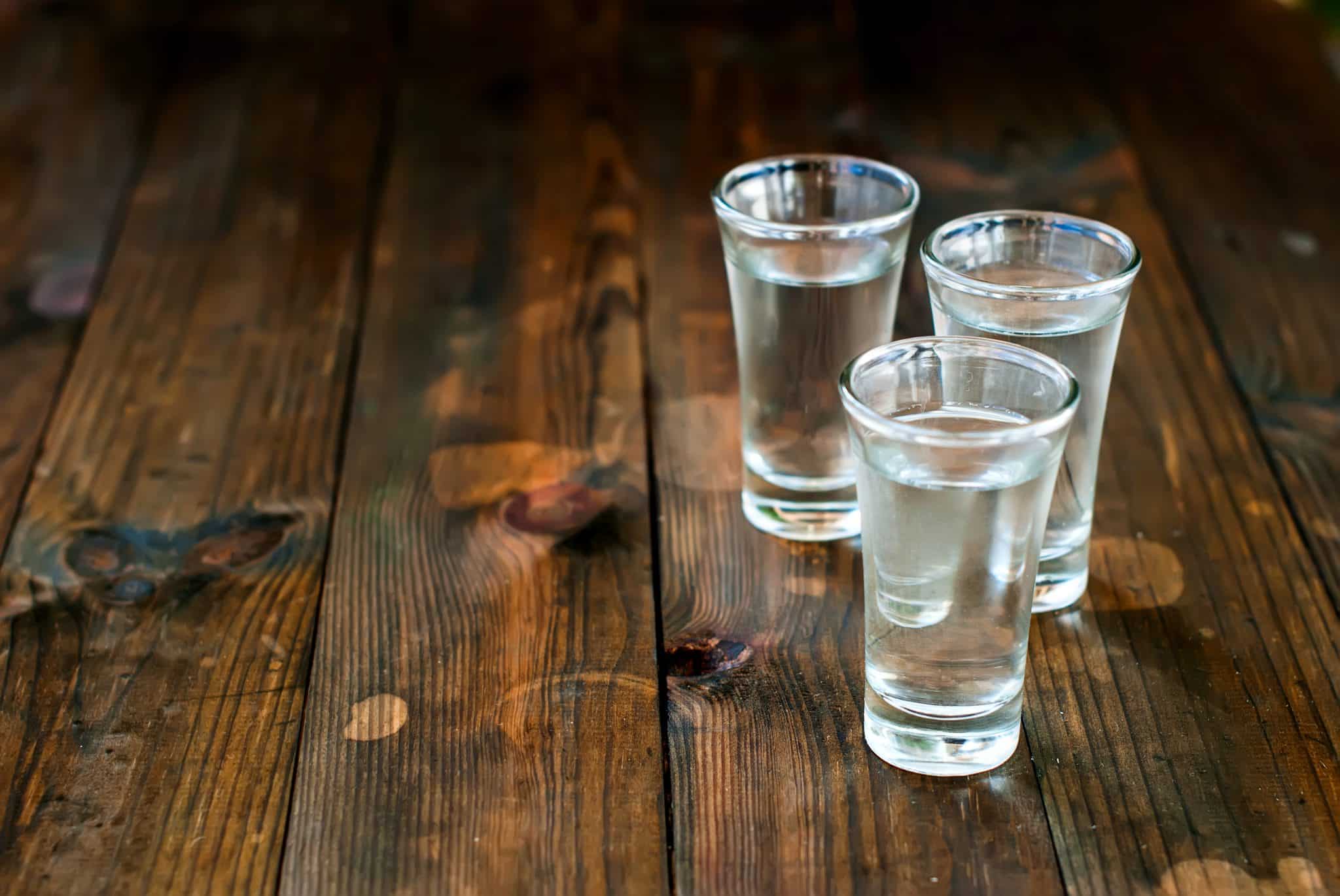 Three small clear glasses of colorless liquid on a rustic wooden table.