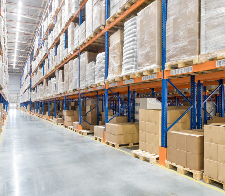 Rows of tall warehouse shelves filled with stacked cardboard boxes and pallets line a wide, clean aisle in a brightly lit industrial storage facility.