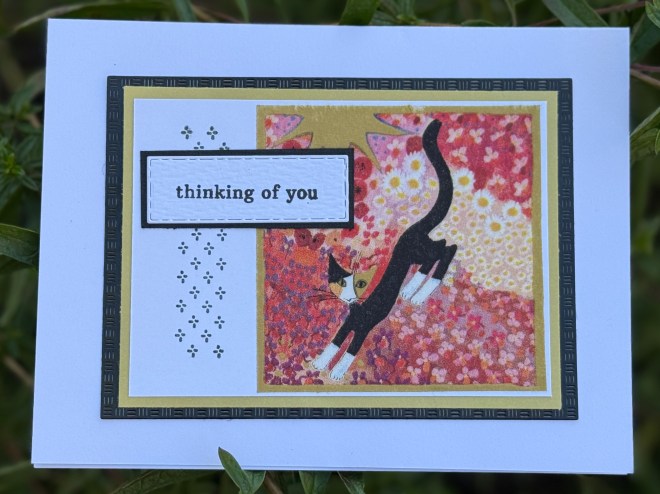 A greeting card featuring a colorful illustration of a black cat with white paws and a yellow background, bordered by decorative edges. The text on the card reads 'thinking of you'.