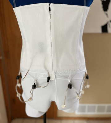 A bodice of a prom dress hanging on a form with fiber optic fabric bundles dangling underneath with some of them installed into a NeoPixel "Dots Strand" strip installed along the waist line