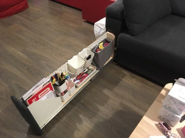 A dark grey couch with a white pegboard on a drawer slide protruding from its arm. The pegboard has a magazine holder, pen holder, and several other miscellaneous bins holding odds and ends on it.