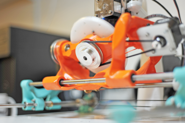 The underside of the rotational base of the Gen5X 3D printer. A belt connects a pulley on the bottom of the stage to a stepper motor on the right side. The carriage for the stage looks organic in nature and is printed in bright orange PLA. The stage can rotate within the carriage which is mounted on two stainless steel rods connected to teal mounting points on either side of the printer (ends of the X-axis).