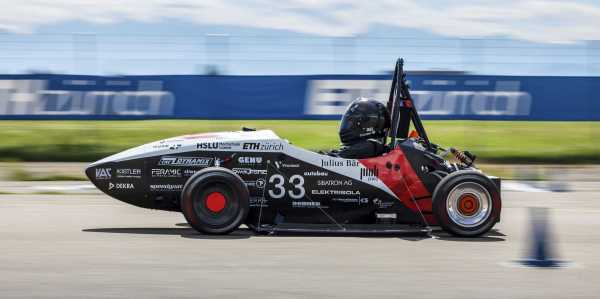 A black race car with white text of sponsors moves across an asphalt surface. There is a blue wall and a green, grassy field in the background. The car has white and red stripes as well.