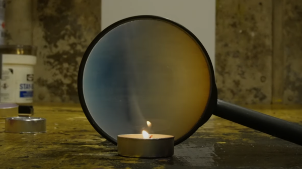 A magnifying glass is seen behind a small tea candle. The magnifying image is projecting the shadow of a column of heated air.