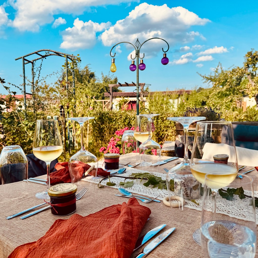 Ein wunderschön eingerichteter Tisch im Freien mit Weingläsern, Utensilien und kleinen Gläsern, umgeben von üppigem Grün und Blumen. Dekorative farbige Lichter hängen an einer nahe gelegenen Struktur unter einem hellblauen Himmel mit Wolken.