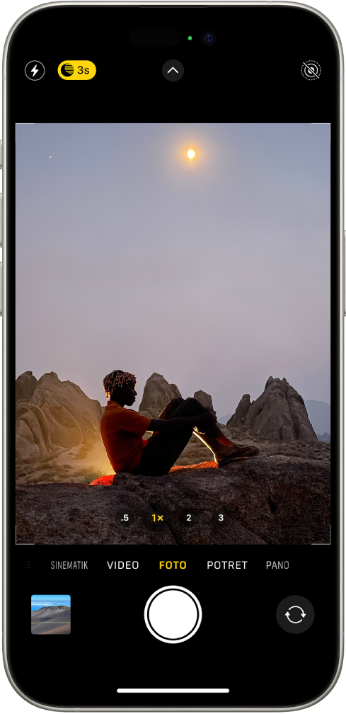 Skrin Kamera dalam mod Foto. Butang untuk denyar dan mod Malam kelihatan di penjuru kiri atas skrin, serta mod Malam diaktifkan. Butang Kawalan Kamera berada di bahagian tengah atas dan butang Live Photo berada di penjuru kanan atas. Di bahagian bawah skrin ialah, dari kiri ke kanan, butang Pemapar Foto dan Video, butang Ambil Gambar dan butang Pemilih Kamera Pandang Belakang. Dalam bingkai kamera, ialah suasana malam.