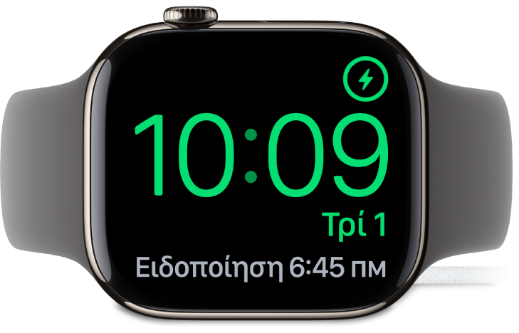 Ένα Apple Watch είναι σε πλάγια θέση και συνδεδεμένο στον φορτιστή του. Στην οθόνη εμφανίζονται το σύμβολο φόρτισης στην πάνω δεξιά γωνία, η τρέχουσα ώρα από κάτω, και η ώρα της επόμενης ειδοποίησης.