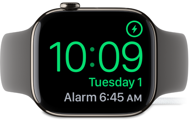 Küljele asetatud ja laadijaga ühendatud Apple Watch, mille ekraanil kuvatakse ülemises paremas nurgas laadimissümbolit, selle all praegust kellaaega ning järgmise äratuse kellaaega.