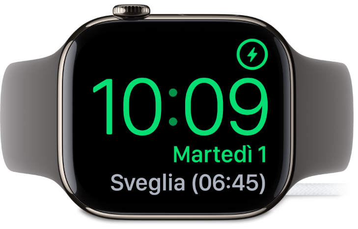 Apple Watch, appoggiato su un lato e collegato al caricabatterie, con lo schermo che mostra il simbolo di ricarica nell’angolo superiore destro, l’ora attuale sotto e l’orario della sveglia successiva.