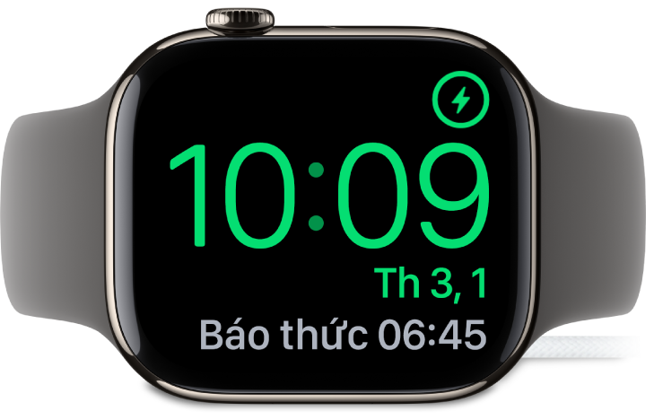 Một Apple Watch được đặt nghiêng và được kết nối với bộ sạc, với màn hình đang hiển thị biểu tượng đang sạc ở góc trên cùng bên phải, thời gian hiện tại ở bên dưới và thời gian của báo thức tiếp theo.