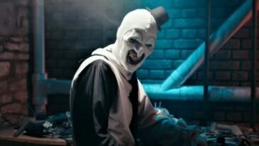 Art the Clown seated at a work bench in a dark basement, smiling in a very unsettling way.