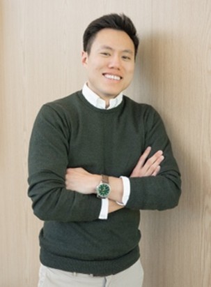 A photo of a man with arms crossed, wearing a dark green sweater over a collared shirt, and a watch, standing against a wooden background.