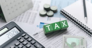 Calculator, tax forms, cash, and blocks spelling “TAX” on a desk