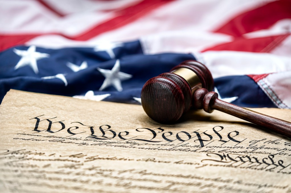 A wooden judge's gavel rests on a copy of the U.S. Constitution, with the opening words 'We the People' visible, set against a folded American flag.