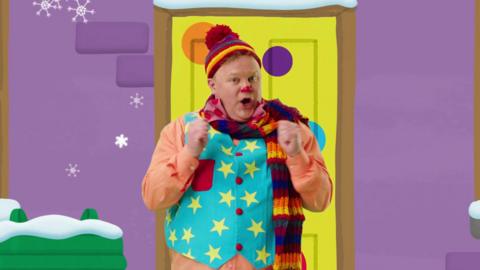Mr Tumble is wearing a hat and scarf standing outside a yellow door.