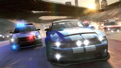 An in-game screenshot of The Crew which shows a blue and white striped rally car racing towards the screen along a concrete river channel, with two police cars and other cars in pursuit.