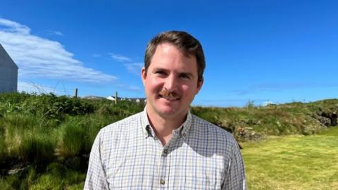 Liam Alasdair Crouse na sheasamh a-muigh agus an eadhar ghorm air a' chùlaibh.