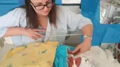Maria et son fils Bennet n'avaient que trois jours. Maria, qui a de longs cheveux bruns et porte des lunettes et un bracelet d'hôpital, est assise, penchée sur un berceau d'hôpital en plastique. Elle tient la petite main de Bennet. Son fils est intubé et est allongé dans le berceau, coiffé d'un petit chapeau, niché dans des couvertures.