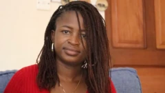 Noella est assise à l'intérieur face à la caméra avec un petit sourire. Elle porte des créoles en argent, un collier et un haut rouge.