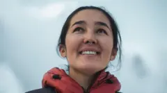 Portrait of River Ahmad during acclimatisation in Nepal before climbing Mount Everest. She is wearing a red insulated mountain jacket, and her black hair is tied back. She is smiling and looking up and to the left, against a backdrop of snow.