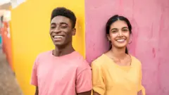 Un jeune homme et une jeune femme se tiennent debout, côte à côte, devant un mur peint de couleurs vives ; le jeune homme porte un t-shirt rose devant une partie jaune du mur, tandis que la jeune femme porte un t-shirt jaune devant une partie rose du mur.
