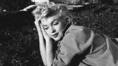 A black and white photo of Marilyn Monroe lying on the ground, with a pensive look and her head resting on her arms.