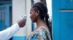Un médecin vérifie la température d'une femme à l'aide d'un thermomètre infrarouge sans contact. Seul le bras du médecin, qui est vêtu de blanc, est montré. La femme qui fait contrôler sa température a les cheveux tressés. Elle porte un haut bleu vif orné d'un motif et se tient à côté d'une porte en bois bleue avec un bâtiment blanc et bleu en arrière-plan.