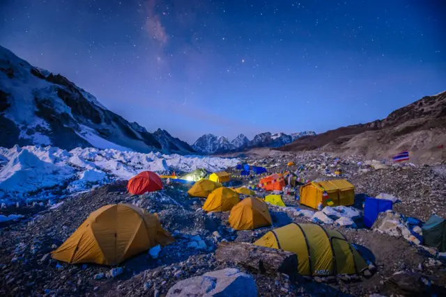 About 10 yellow and orange tents pitched on rocky scree, amid snow covered mountains, at the Everest Base Camp, pictured in low light at dusk or dawn,